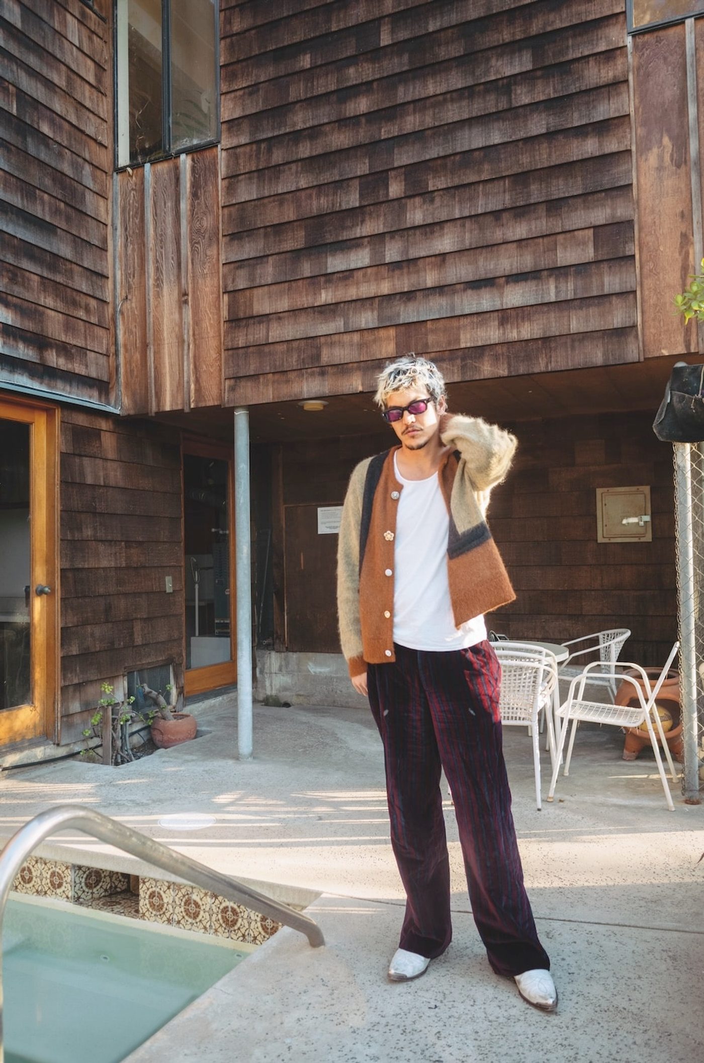 Anthony Cataldo standing beside a swimming pool in front of a cedar-shingled building, bleached hair and red-tinted sunglasses, wearing a colour-blocked mohair cardigan over a white vest, dark red striped velvet trousers and white cowboy boots