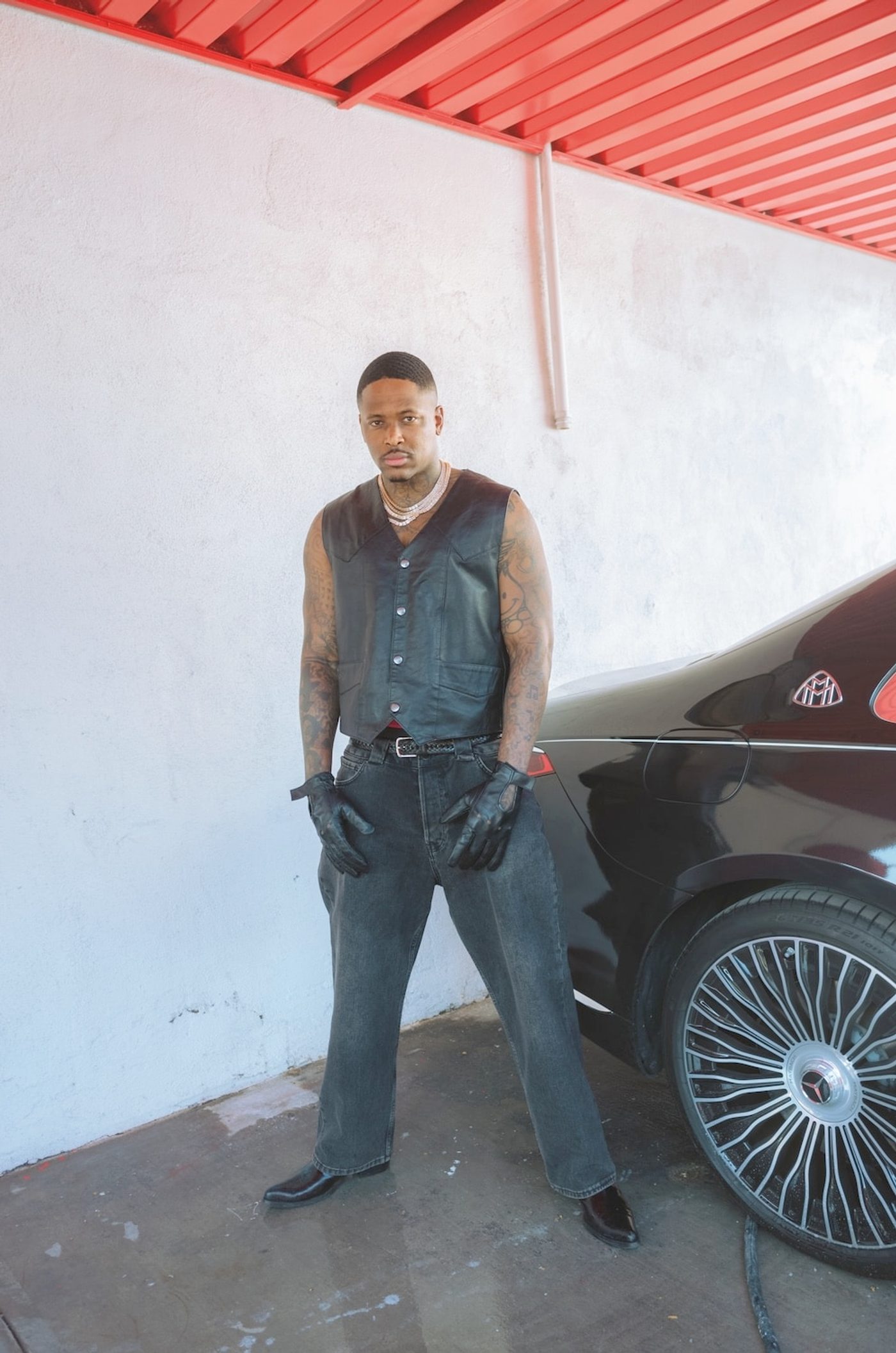 YG standing against a white stucco wall in a black leather waistcoat, leather gloves and loose grey jeans, tattooed arms and layered chains, a black Maybach parked beside him under a red steel awning